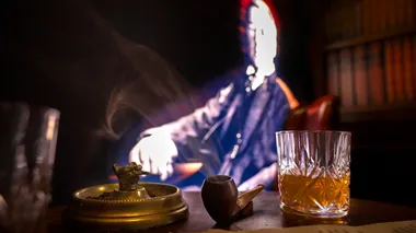 A smoking pipe, ash tray and a whiskey glass on a table in front of a projection of Winston Churchill in a chair.