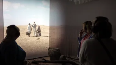 Silhouette of a visitor in front of a projection on the wall, where three actors in simple clothes walk in a dune landscape under a blue sky.