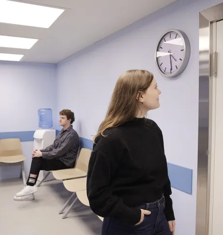 Inside the waiting room, a woman looks at the clock while a man sits down on a waiting bench.