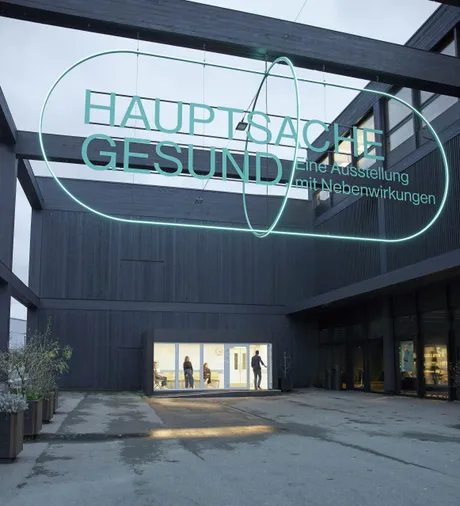 View of the exterior, where a big abstract pill hangs from the construction with the title of the exhibition inside.
