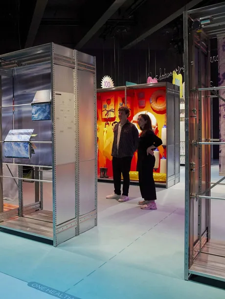 Two people stand in between rows of big showcases, an abstract and minimal design resembling a supermarket layout.