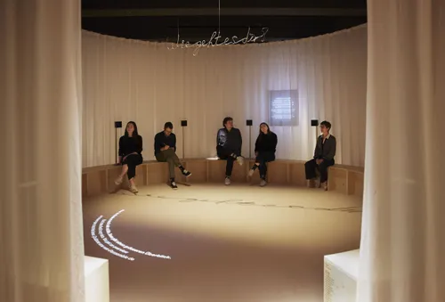 People sit in a circular space on a bench between curtains. Above them hangs a question, 'How are you?', bent in iron. A small text is projected on the floor.