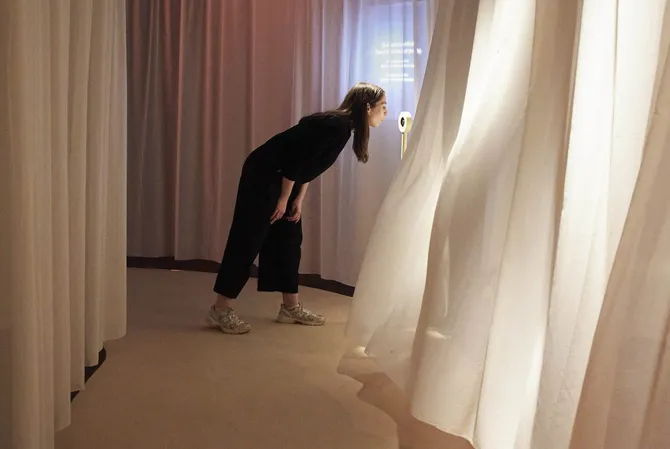 A woman stands in a pathway of curtains on both sides. She blows on an interactive on the right. When she does that, wind blows the curtains upward on the right.