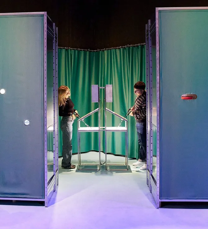 A sightline into a space with green medical curtains, where two people on opposite sides use a physical interactive that resembles an operation. Above them hangs a sentence bent from iron that says 'Was stent dir zu?'.