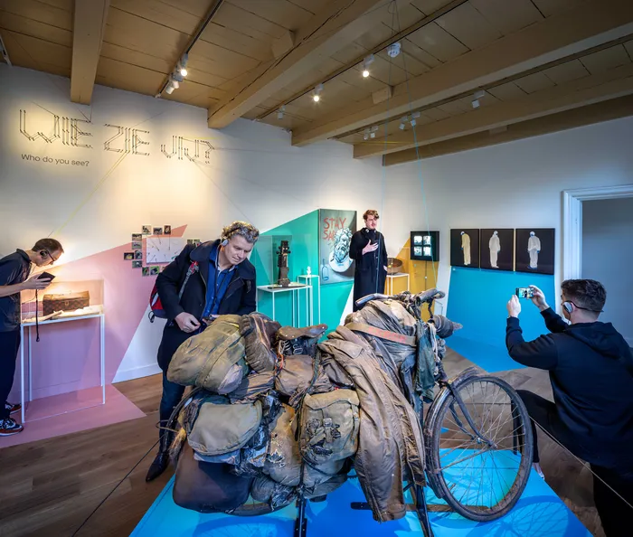 A packed bike of a person experiencing homelessness stands in the center of the room. Colourful paint on the walls frame the stories presented there.