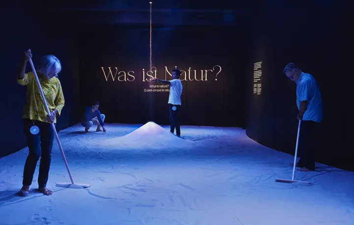 Three people stand in a black room, with the floor covered in almost white sand. The room has a blue hue to it. On the back wall it says 'What is nature?'. Two people use levelling rakes to make shapes in the sand. Another person stands in the middle, where sand falls from the ceiling into a big pile. He has his arm outstretched to feel the falling sand.