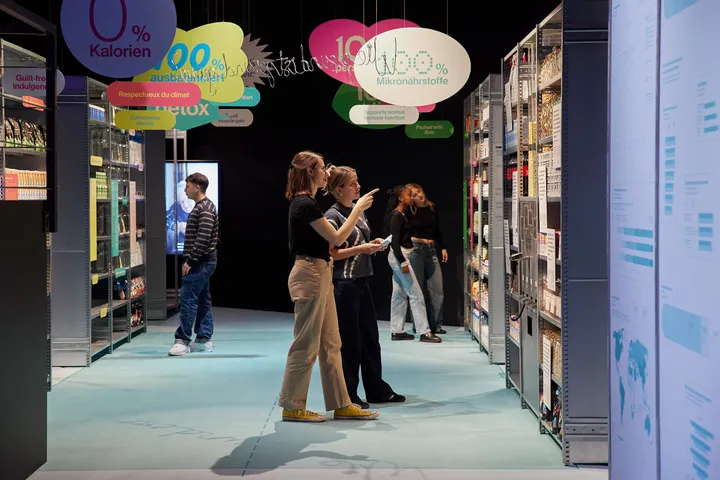 Two women stand in a space that resembles and abstract supermarket, with all different graphic advertisement expressions above them.
