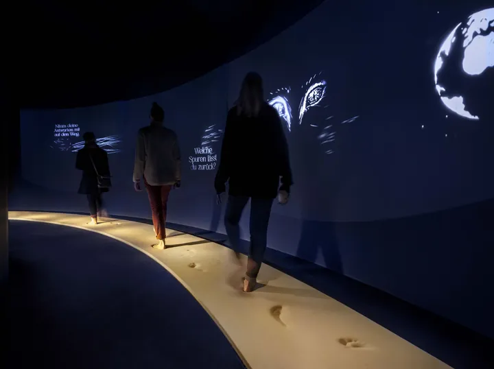 Three visitors walk over a curved foam pathway that looks like sand, while projections follow them on the wall next to them.