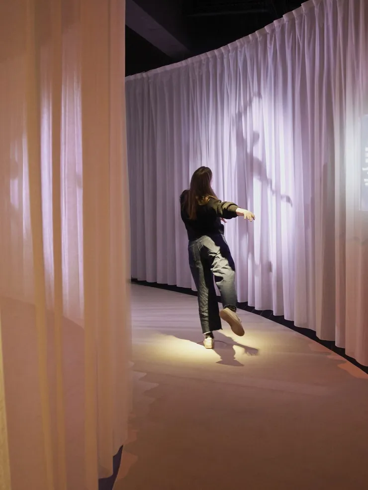 In a pathway between warm beige curtains, a woman dances in an elegant move that is mirrored in silhouette on the curtain.