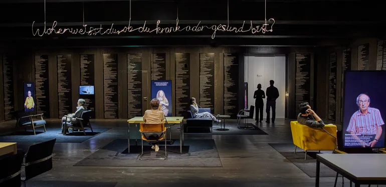 A wide shot of a room where different tables and chairs form places to sit down opposite a video portrait. The walls are covered in small text of possible diagnoses.