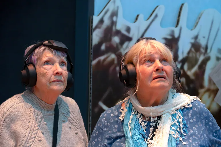 Two visitors listening to their audio tour look up with expressions ranging from shock to bewilderment.