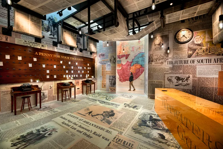 Overview image of the last space of the exhibition. The walls and floors are covered in newspaper, and in the corner stand three tables with typewriters. Above them papers hang on a wooden board, engraved with the text 'A penny for your thoughts'. Above visitors heads, a monumental printing press installation turns slowly.