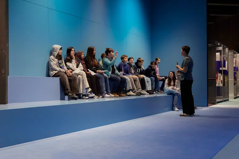 A school group sitting on broad steps that serve as group seating, with a tour guide standing in front of them.