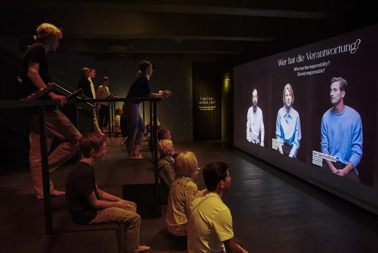 People sit on a big staircase, where upstairs, people lean over the railing. They look to a wall on the right, where three figures in a larger-than-life projection share their opinion about the question 'Who shares in the responsibility?' that fills the screen above them.