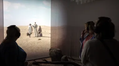 Silhouette of a visitor in front of a projection on the wall, where three actors in simple clothes walk in a dune landscape under a blue sky.