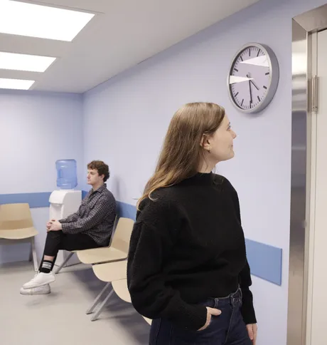 Inside the waiting room, a woman looks at the clock while a man sits down on a waiting bench.