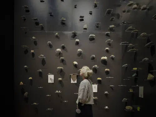 An older woman looks up to a black wall filled with seemingly floating stones on display.