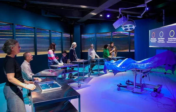 People stand at different iron desks around an operating chair, with screens behind the chair posing different dilemmas. On their iron desk are three buttons on a medical tool plate, to give your solution to the dilemma.