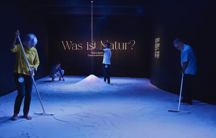 Three people stand in a black room, with the floor covered in almost white sand. The room has a blue hue to it. On the back wall it says 'What is nature?'. Two people use levelling rakes to make shapes in the sand. Another person stands in the middle, where sand falls from the ceiling into a big pile. He has his arm outstretched to feel the falling sand.