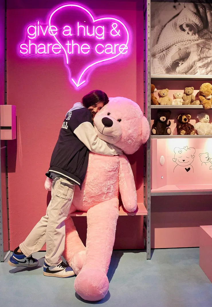 A girl hugging a life-sized teddy bear under a pink neon sign that says 'give a hug and share the care'.