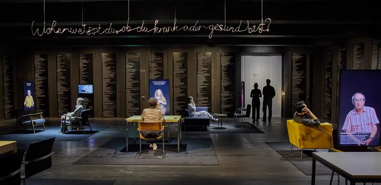 A wide shot of a room where different tables and chairs form places to sit down opposite a video portrait. The walls are covered in small text of possible diagnoses.